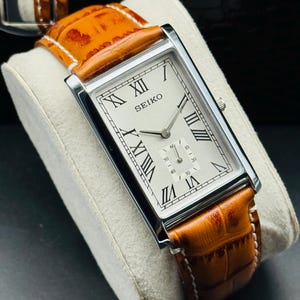 Op de afbeelding: Een rechthoekig Seiko horloge met een zilveren kast en een cr&egrave;mekleurige wijzerplaat. Het horloge heeft zwarte Romeinse cijfers en een bruine leren band. De band is ongeveer 2 cm breed. Het horloge wordt getoond op een wit kussen.