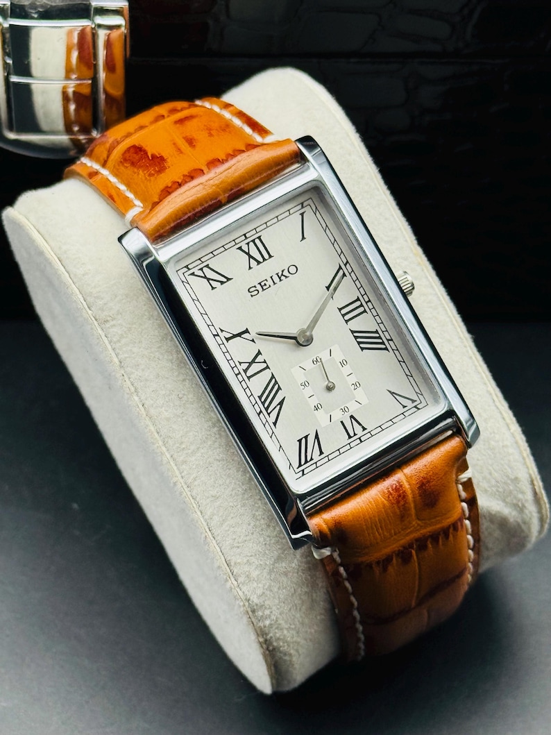 Op de afbeelding: Een rechthoekig Seiko horloge met een zilveren kast en een cr&egrave;mekleurige wijzerplaat. Het horloge heeft zwarte Romeinse cijfers en een bruine leren band. De band is ongeveer 2 cm breed. Het horloge wordt getoond op een wit kussen.