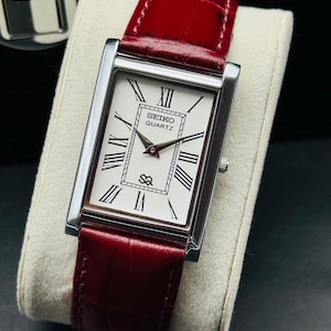 May include: A rectangular Seiko Quartz watch with a silver case and a white face featuring black Roman numerals. The watch has a red leather band. The watch is displayed on a white cushion.