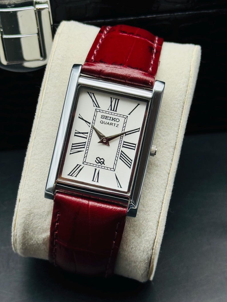 May include: A rectangular Seiko Quartz watch with a silver case and a white face featuring black Roman numerals. The watch has a red leather band. The watch is displayed on a white cushion.
