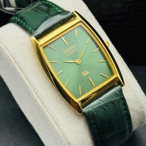 May include: A gold-toned Seiko Quartz watch with a rectangular green face and gold hands. The watch has a green leather band. The watch is displayed on a white surface.