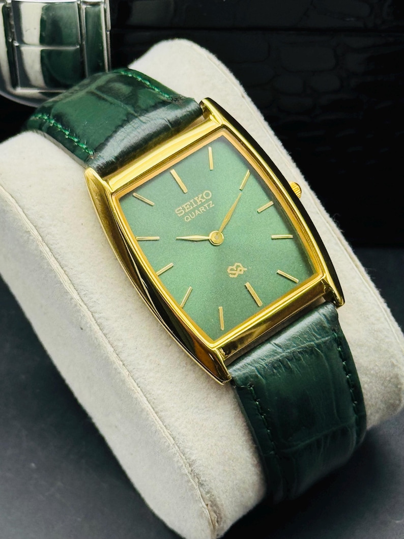 May include: A gold-toned Seiko Quartz watch with a rectangular green face and gold hands. The watch has a green leather band. The watch is displayed on a white surface.