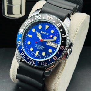Könnte beinhalten: Eine Seiko Marine Master automatische Profi-Uhr mit blauem Zifferblatt und schwarzer Lünette. Die Uhr hat ein schwarzes Kautschukarmband und eine Datumsanzeige. Die Uhr wird auf einem weißen Kissen präsentiert.