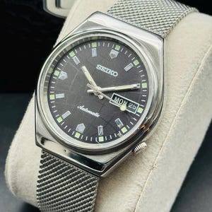 Puede incluir: Un reloj de pulsera automático Seiko plateado con esfera gris oscuro y correa de malla. La esfera del reloj muestra el día y la fecha, con marcadores y agujas luminosas. El reloj se muestra sobre un cojín de color claro.