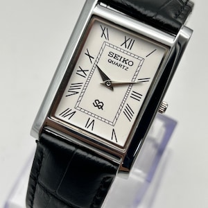 Puede incluir: Un reloj Seiko Quartz rectangular con esfera blanca, n&uacute;meros romanos negros y manecillas negras. El reloj tiene una caja plateada y una correa de cuero negro. La esfera muestra el logotipo de Seiko y la palabra "QUARTZ".