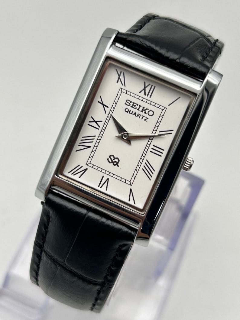 Puede incluir: Un reloj Seiko Quartz rectangular con esfera blanca, n&uacute;meros romanos negros y manecillas negras. El reloj tiene una caja plateada y una correa de cuero negro. La esfera muestra el logotipo de Seiko y la palabra "QUARTZ".