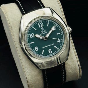 May include: A silver watch with a dark green face and black leather strap. The watch face has white numerals and hands, with the text "TUDOR OYSTER" and "SHOCK RESISTING". The watch is presented on a cream-coloured cushion.