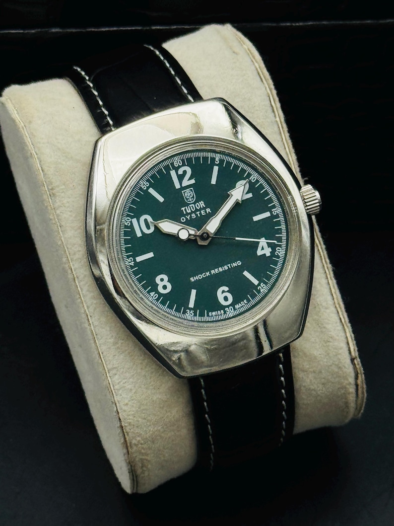May include: A silver watch with a dark green face and black leather strap. The watch face has white numerals and hands, with the text "TUDOR OYSTER" and "SHOCK RESISTING". The watch is presented on a cream-coloured cushion.