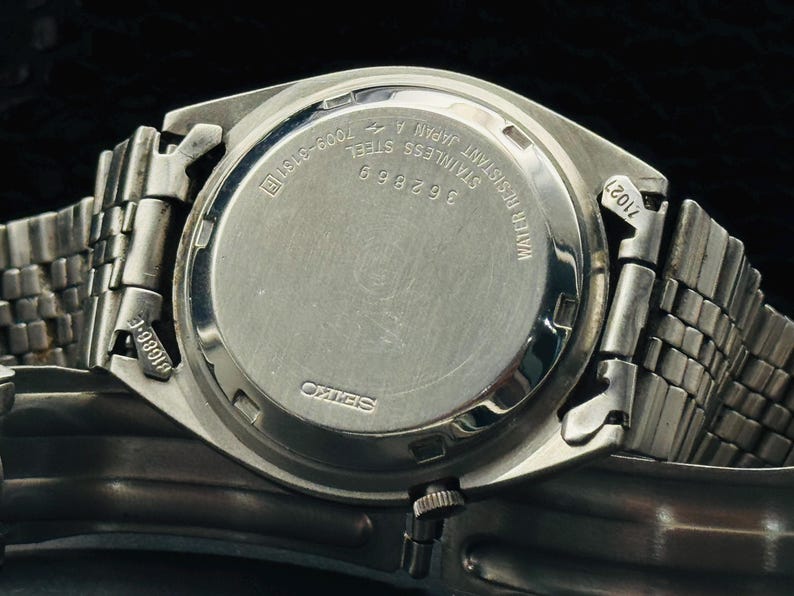 May include: Close-up of a stainless steel Seiko watch. The watch back features the text "WATER RESISTANT JAPAN 4 STAINLESS STEEL 362869 7009-3131R". The watch band is a silver-toned metal.