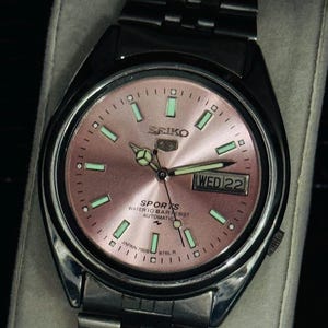 May include: A stainless steel Seiko Sports automatic wristwatch with a pink dial. The watch face features luminous green markers and hands, a day-date display, and the text "SEIKO SPORTS WATER 10 BAR RESIST AUTOMATIC". The watch band is stainless steel.
