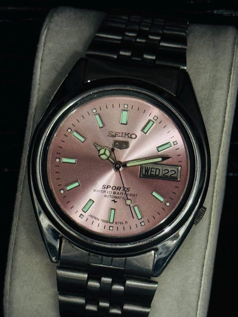 May include: A stainless steel Seiko Sports automatic wristwatch with a pink dial. The watch face features luminous green markers and hands, a day-date display, and the text "SEIKO SPORTS WATER 10 BAR RESIST AUTOMATIC". The watch band is stainless steel.