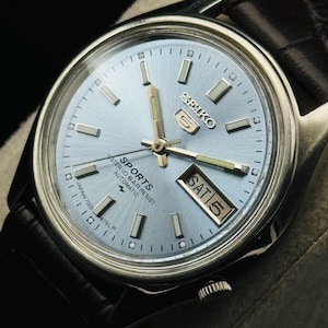 May include: A silver-toned Seiko 5 Sports watch with a light blue face and black leather band. The watch face displays the day and date, with the day reading "SAT 5". The watch has silver hour markers and hands.