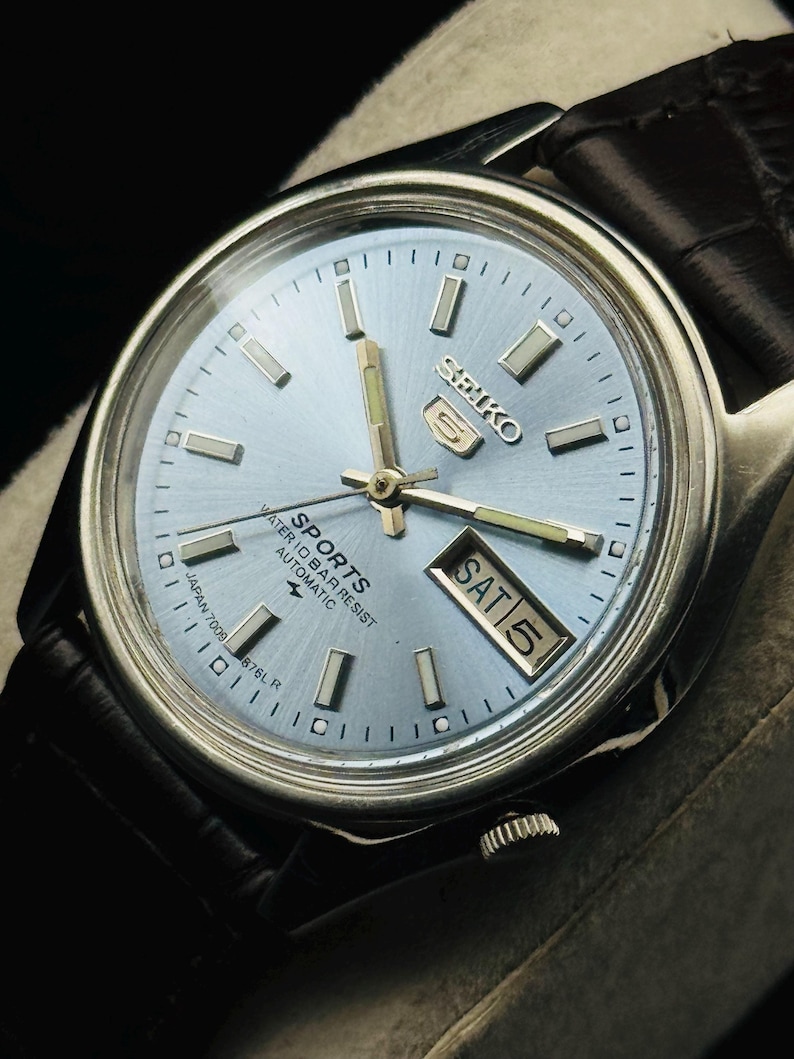May include: A silver-toned Seiko 5 Sports watch with a light blue face and black leather band. The watch face displays the day and date, with the day reading "SAT 5". The watch has silver hour markers and hands.