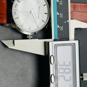 Puede incluir: Un reloj Jaeger-LeCoultre Club plateado con esfera blanca y agujas plateadas, con una correa de cuero marr&oacute;n. El reloj se mide con un calibrador digital, mostrando una medida de 3,827 cm.
