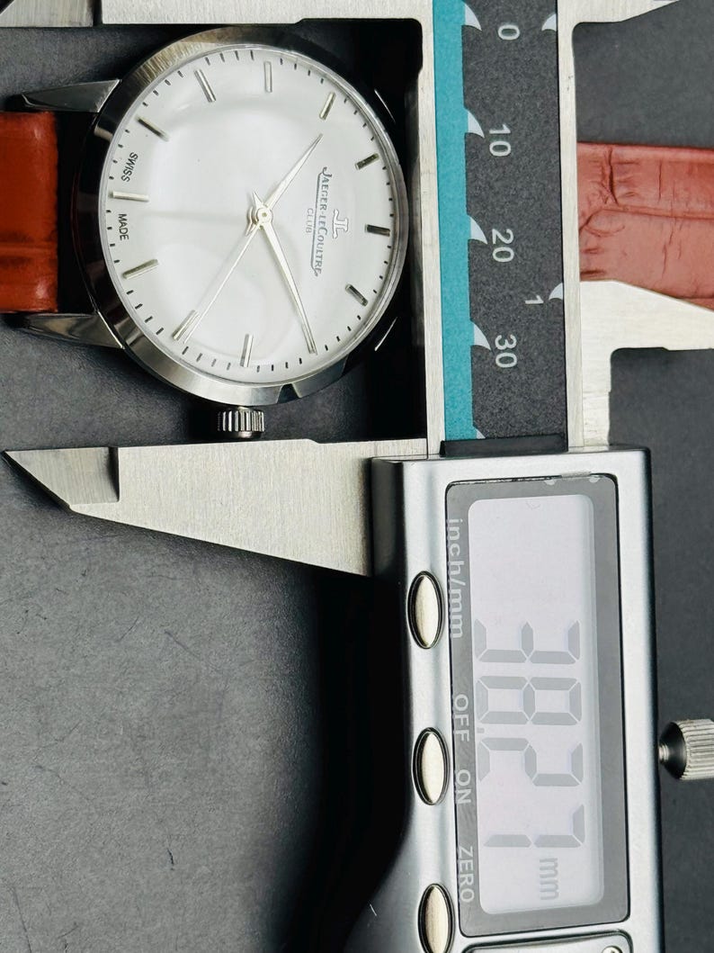 Puede incluir: Un reloj Jaeger-LeCoultre Club plateado con esfera blanca y agujas plateadas, con una correa de cuero marr&oacute;n. El reloj se mide con un calibrador digital, mostrando una medida de 3,827 cm.