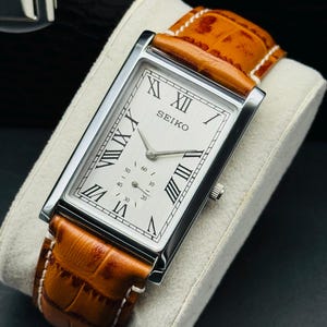 Könnte beinhalten: Eine rechteckige Seiko-Uhr mit silbernem Gehäuse und weißem Zifferblatt mit schwarzen römischen Ziffern. Die Uhr hat ein braunes Lederarmband mit weißen Nähten. Die Uhr wird auf einem grauen Kissen präsentiert.