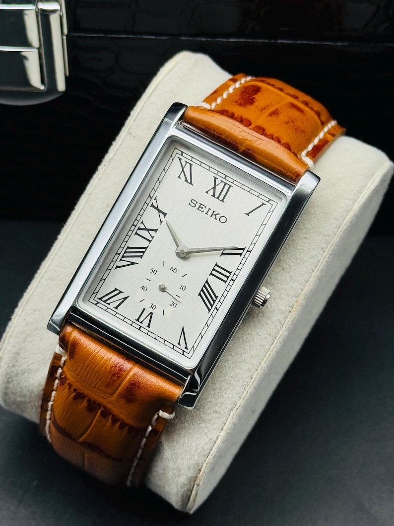 Op de afbeelding: Een rechthoekig Seiko horloge met een zilveren kast en een witte wijzerplaat met zwarte Romeinse cijfers. Het horloge heeft een bruine leren band met witte stiksels. Het horloge wordt getoond op een grijs kussen.
