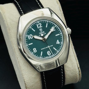 May include: A silver wristwatch with a dark green face and a black leather strap. The watch face displays white numerals and hands, with the brand name "TUDOR". The strap is black with white stitching.