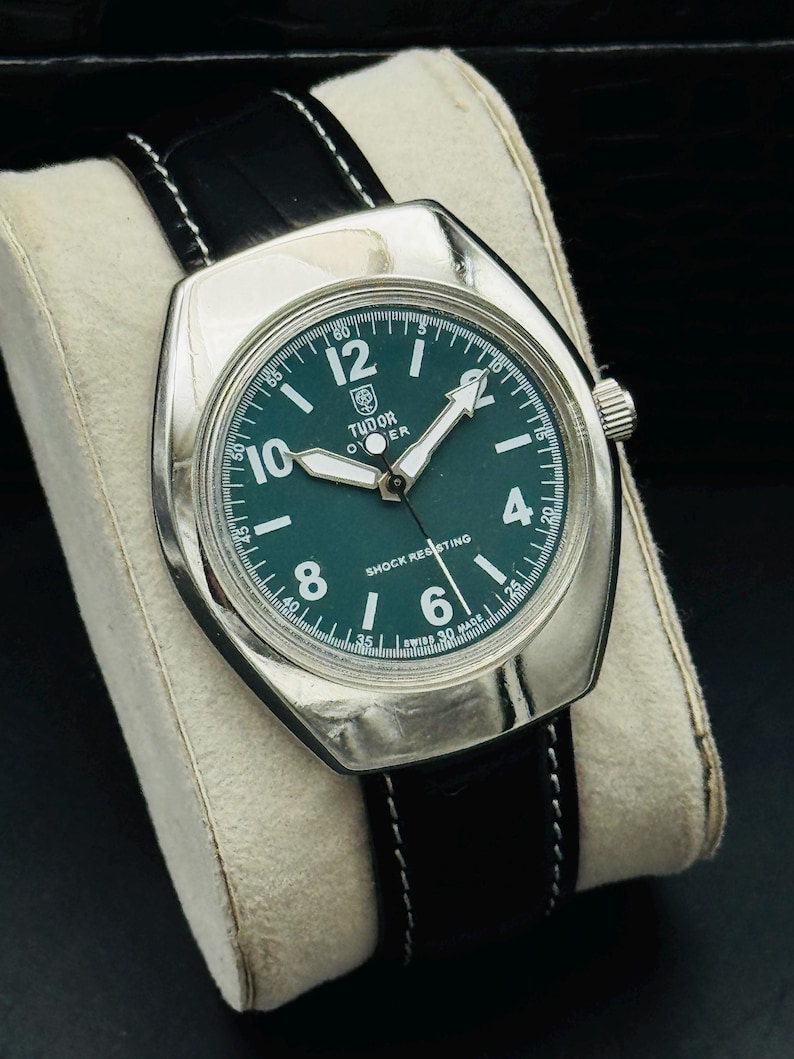 May include: A silver wristwatch with a dark green face and a black leather strap. The watch face displays white numerals and hands, with the brand name "TUDOR". The strap is black with white stitching.