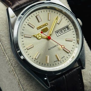 May include: Silver-toned Seiko automatic watch with a light-colored face, gold hands, and a brown leather strap. The watch face displays the day and date, with the text "SEIKO" and "AUTOMATIC". The watch is marked "JAPAN MADE".