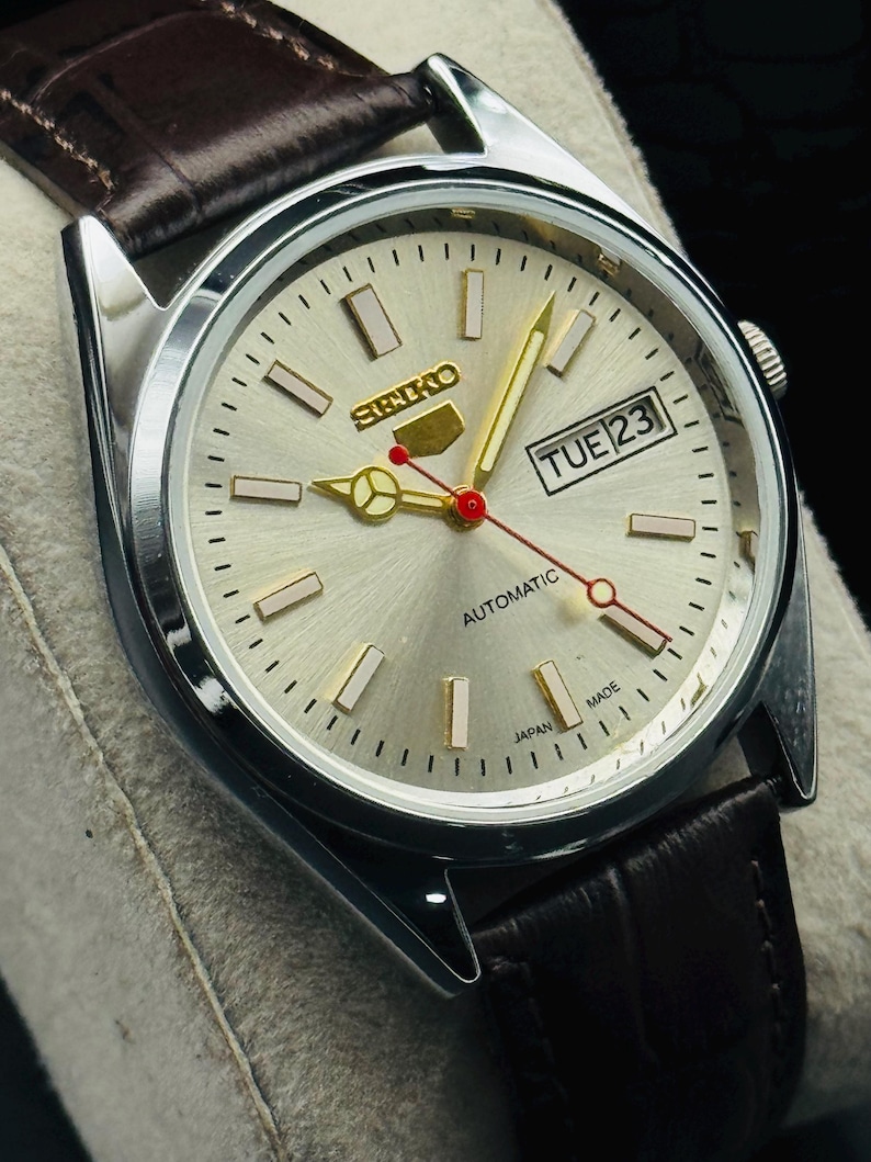 May include: Silver-toned Seiko automatic watch with a light-colored face, gold hands, and a brown leather strap. The watch face displays the day and date, with the text "SEIKO" and "AUTOMATIC". The watch is marked "JAPAN MADE".