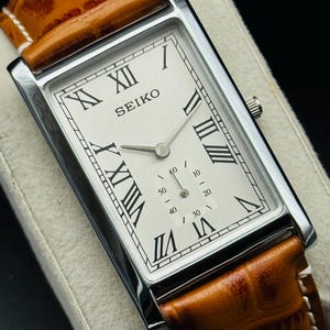 Op de afbeelding: Een rechthoekig Seiko horloge met een zilveren kast en een lichtgekleurde wijzerplaat. Het horloge heeft Romeinse cijfers als uurmarkeringen en een bruine leren band. De wijzerplaat toont de merknaam "SEIKO".