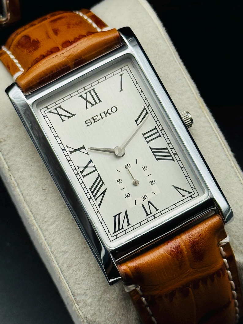 Op de afbeelding: Een rechthoekig Seiko horloge met een zilveren kast en een lichtgekleurde wijzerplaat. Het horloge heeft Romeinse cijfers als uurmarkeringen en een bruine leren band. De wijzerplaat toont de merknaam "SEIKO".