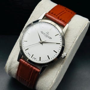 Puede incluir: Un reloj Jaeger-LeCoultre Club plateado con esfera blanca y marcadores negros. Tiene una correa de cuero marr&oacute;n. El reloj se exhibe sobre un coj&iacute;n gris. Fabricado en Suiza.