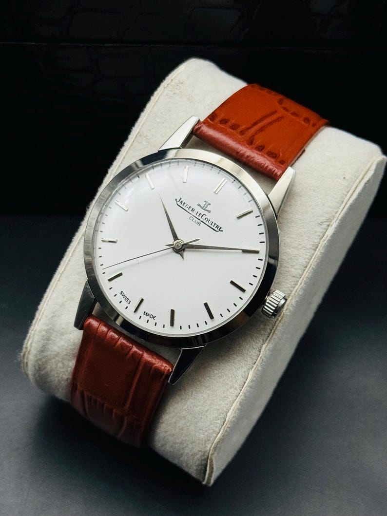 Puede incluir: Un reloj Jaeger-LeCoultre Club plateado con esfera blanca y marcadores negros. Tiene una correa de cuero marr&oacute;n. El reloj se exhibe sobre un coj&iacute;n gris. Fabricado en Suiza.