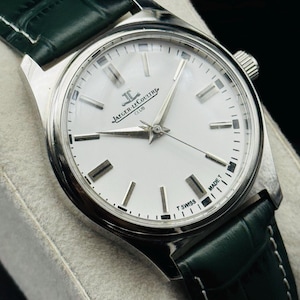 May include: A silver-toned Jaeger-LeCoultre Club watch with a white face and a dark green leather strap. The watch has silver hands and markers, and the face reads "T SWISS MADE T".