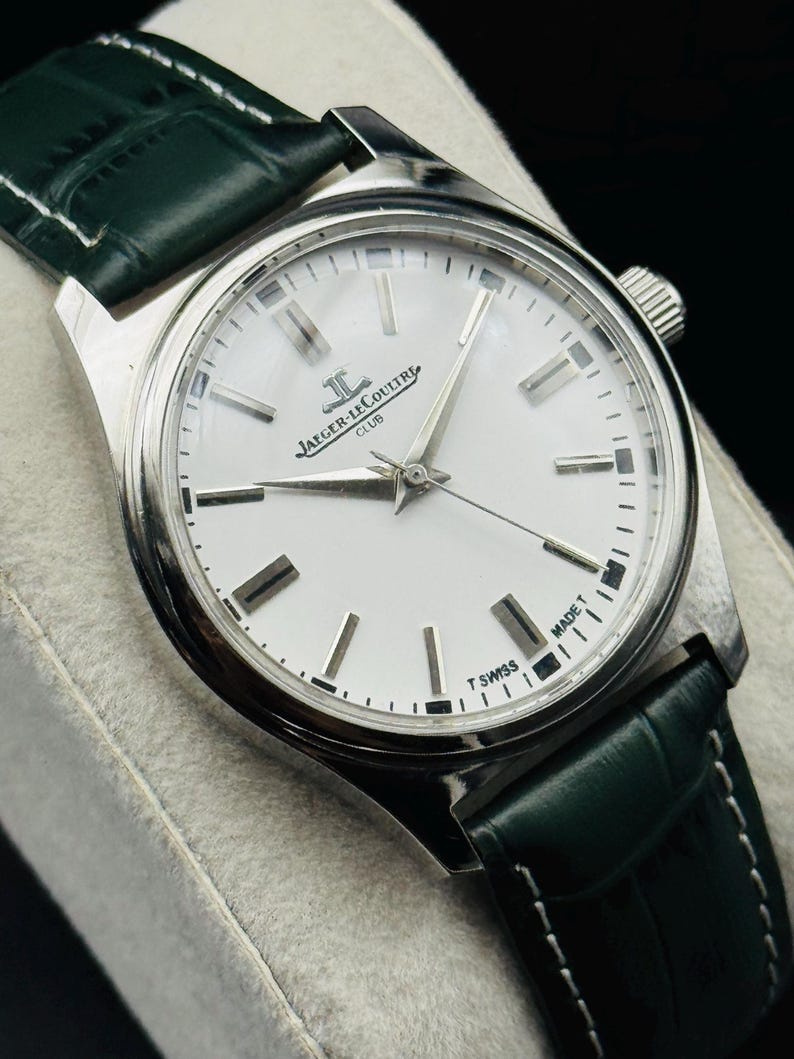 May include: A silver-toned Jaeger-LeCoultre Club watch with a white face and a dark green leather strap. The watch has silver hands and markers, and the face reads "T SWISS MADE T".