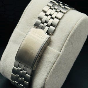 May include: A close-up of a stainless steel watch band with a clasp. The band is composed of rectangular links and is resting on a white, plush surface. The clasp is engraved with the word "SEIKO".