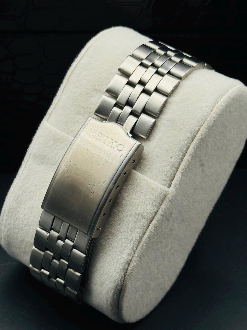 May include: A close-up of a stainless steel watch band with a clasp. The band is composed of rectangular links and is resting on a white, plush surface. The clasp is engraved with the word "SEIKO".