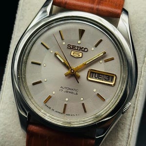 May include: A silver Seiko 5 automatic watch with a light grey face, gold hands and markers, and a brown leather strap. The watch face displays the day and date, and the text "AUTOMATIC 17 JEWELS" and "MADE IN JAPAN".