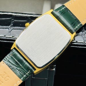 May include: Close-up of a gold-toned rectangular watch with a silver face and green leather strap. The watch has a gold bezel and a green leather strap. The watch is on a black surface.