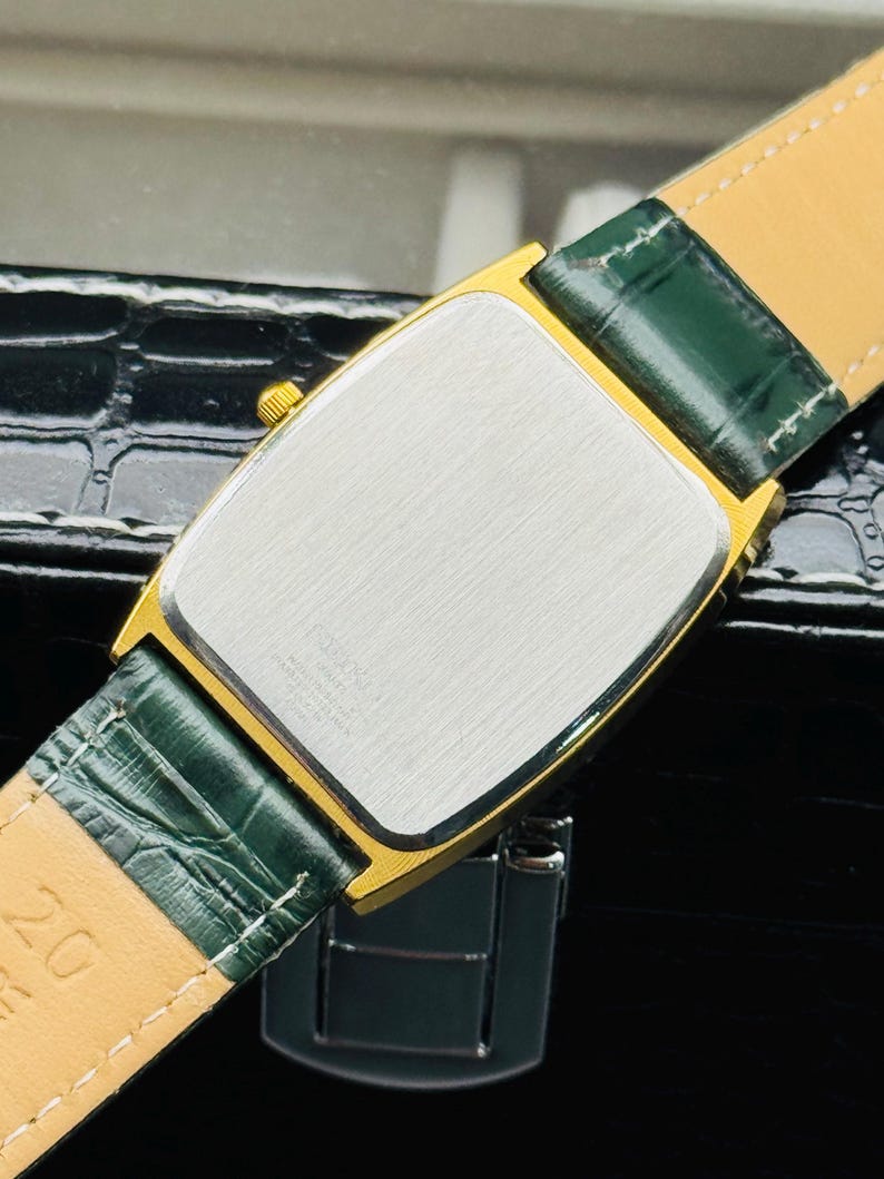 May include: Close-up of a gold-toned rectangular watch with a silver face and green leather strap. The watch has a gold bezel and a green leather strap. The watch is on a black surface.