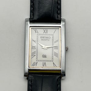 Puede incluir: Un reloj Seiko Quartz rectangular con caja plateada y correa de cuero negro. La esfera blanca presenta n&uacute;meros romanos, detalles dorados y el logotipo de Seiko. El reloj tiene un dise&ntilde;o cl&aacute;sico y elegante.