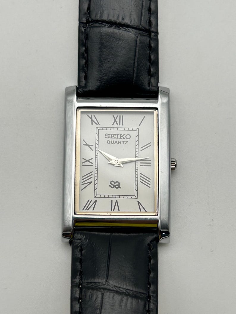 Puede incluir: Un reloj Seiko Quartz rectangular con caja plateada y correa de cuero negro. La esfera blanca presenta n&uacute;meros romanos, detalles dorados y el logotipo de Seiko. El reloj tiene un dise&ntilde;o cl&aacute;sico y elegante.