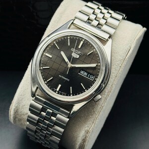 Op de afbeelding: Een zilverkleurig Seiko 5 automatisch horloge met een zwarte wijzerplaat en een roestvrijstalen band. De wijzerplaat toont de dag en de datum. Het horloge ligt op een wit kussen.