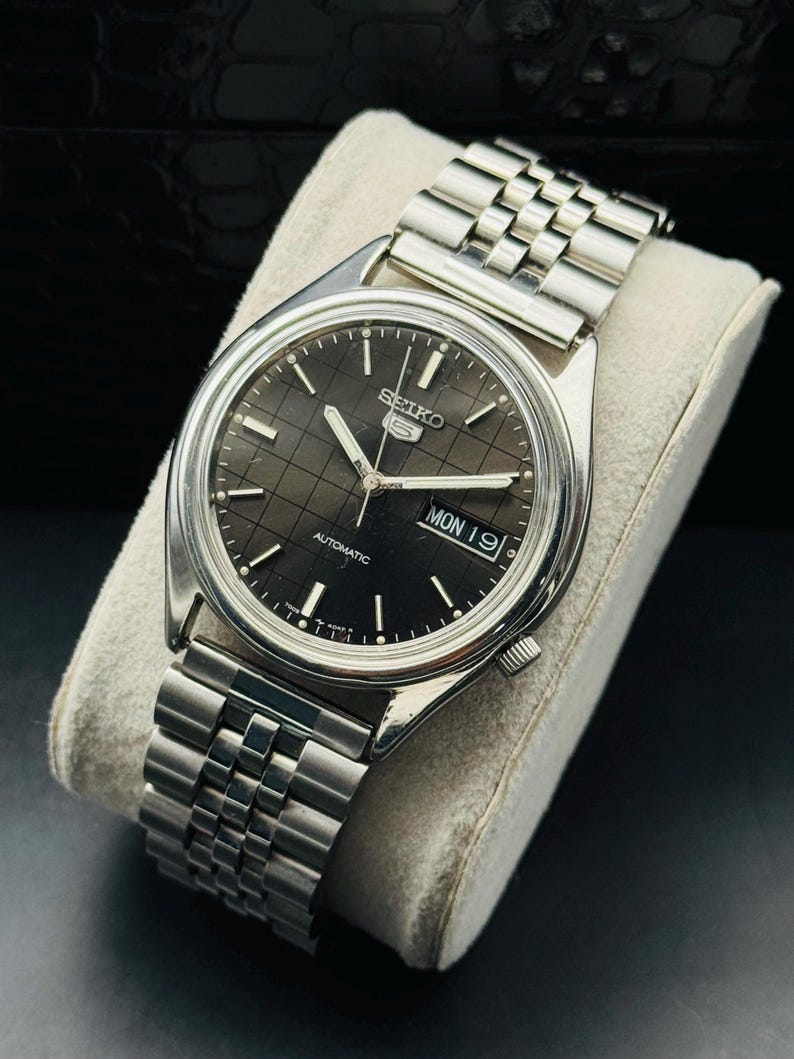 Op de afbeelding: Een zilverkleurig Seiko 5 automatisch horloge met een zwarte wijzerplaat en een roestvrijstalen band. De wijzerplaat toont de dag en de datum. Het horloge ligt op een wit kussen.