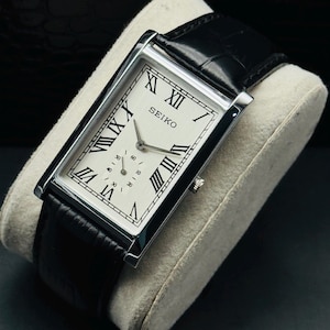 May include: A rectangular Seiko watch with a silver case and a white face featuring black Roman numerals. The watch has a small seconds dial and is set on a light grey cushion. The black leather strap is visible.