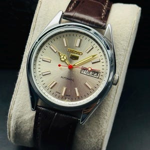 May include: Silver-toned Seiko automatic watch with a light-colored face, gold hands, and a red second hand. The watch features a day-date display and a brown leather strap. The watch is displayed on a light-colored cushion.