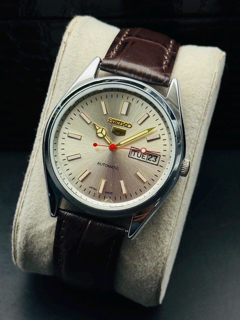 May include: Silver-toned Seiko automatic watch with a light-colored face, gold hands, and a red second hand. The watch features a day-date display and a brown leather strap. The watch is displayed on a light-colored cushion.