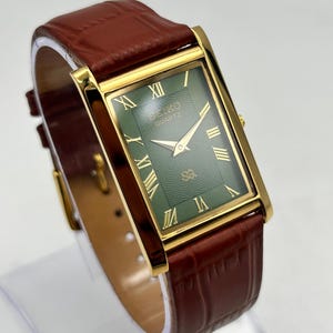 Puede incluir: Un reloj Seiko Quartz rectangular con caja dorada y esfera verde. El reloj presenta n&uacute;meros romanos y agujas doradas, y una correa de cuero marr&oacute;n. El reloj se exhibe sobre un soporte transparente.