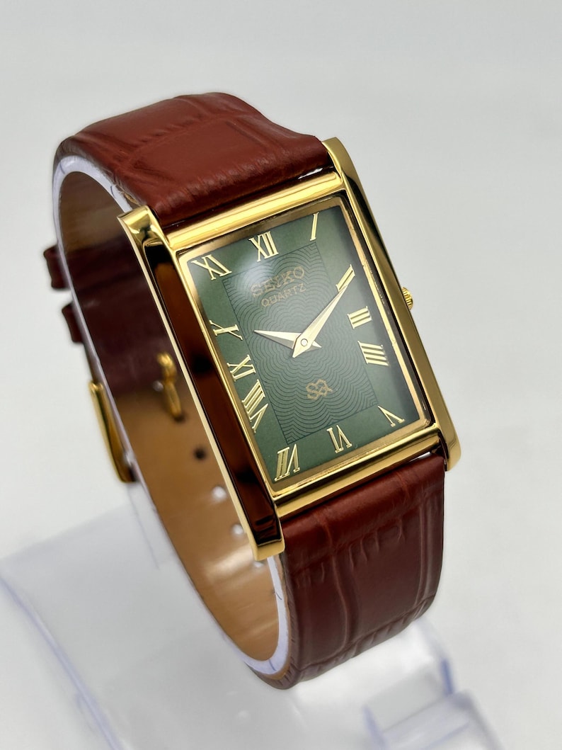 Puede incluir: Un reloj Seiko Quartz rectangular con caja dorada y esfera verde. El reloj presenta n&uacute;meros romanos y agujas doradas, y una correa de cuero marr&oacute;n. El reloj se exhibe sobre un soporte transparente.