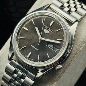 Op de afbeelding: Een zilverkleurig Seiko 5 automatisch horloge met een donkergrijze geruite wijzerplaat. Het horloge heeft een dag- en datumweergave en een zilveren schakelarmband. Het horloge wordt gepresenteerd op een lichtgrijze ondergrond.