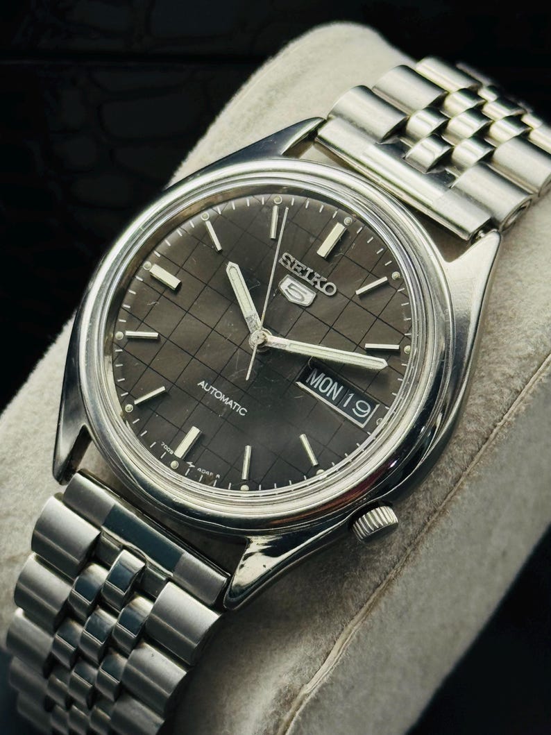 Op de afbeelding: Een zilverkleurig Seiko 5 automatisch horloge met een donkergrijze geruite wijzerplaat. Het horloge heeft een dag- en datumweergave en een zilveren schakelarmband. Het horloge wordt gepresenteerd op een lichtgrijze ondergrond.