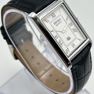 Puede incluir: Un reloj Seiko Quartz rectangular con esfera blanca y n&uacute;meros romanos negros. El reloj tiene una caja plateada y una correa de cuero negro. La esfera muestra el logotipo de Seiko y un peque&ntilde;o emblema. La correa mide aproximadamente 20 cm.
