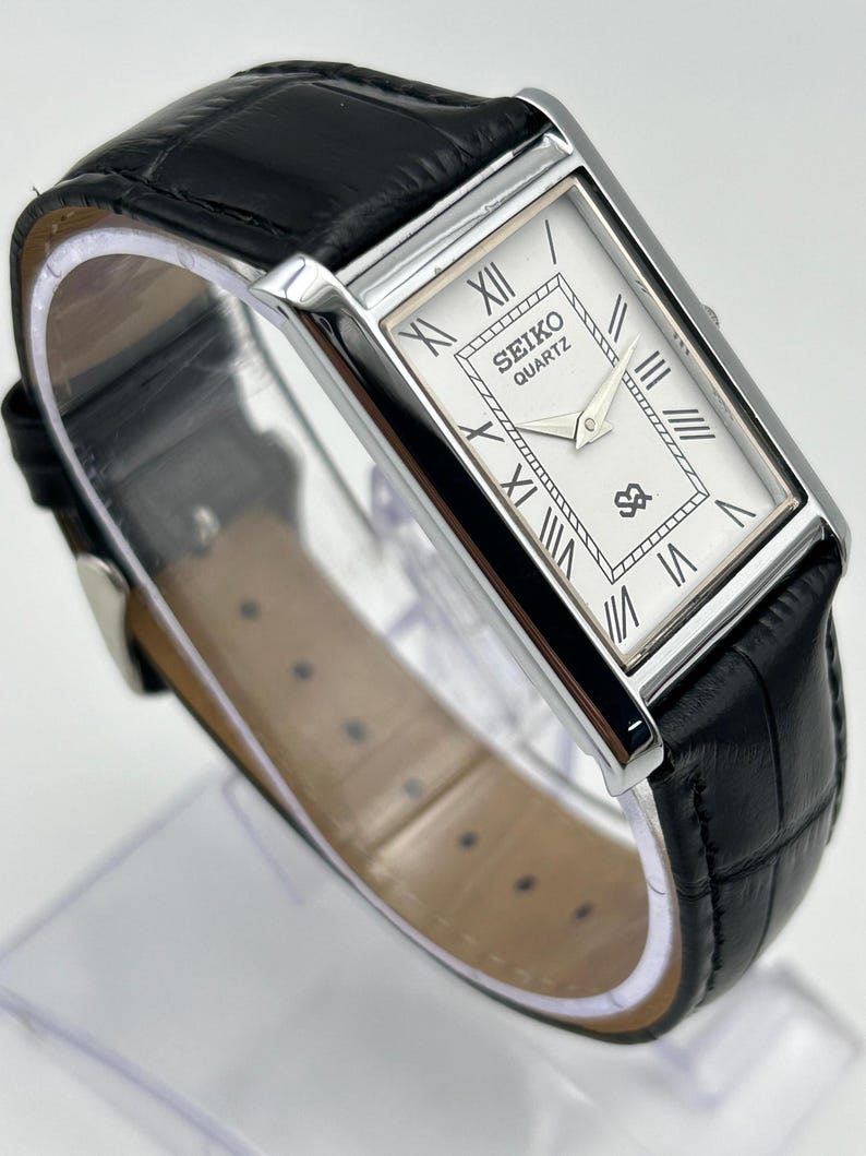 Puede incluir: Un reloj Seiko Quartz rectangular con esfera blanca y n&uacute;meros romanos negros. El reloj tiene una caja plateada y una correa de cuero negro. La esfera muestra el logotipo de Seiko y un peque&ntilde;o emblema. La correa mide aproximadamente 20 cm.