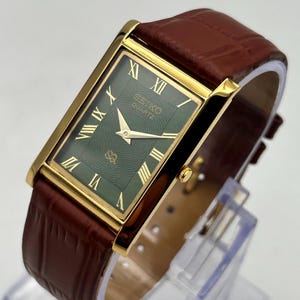 Puede incluir: Un reloj Seiko Quartz rectangular con caja dorada y esfera verde con n&uacute;meros romanos. El reloj tiene una correa de cuero marr&oacute;n. El reloj se muestra sobre un soporte transparente.